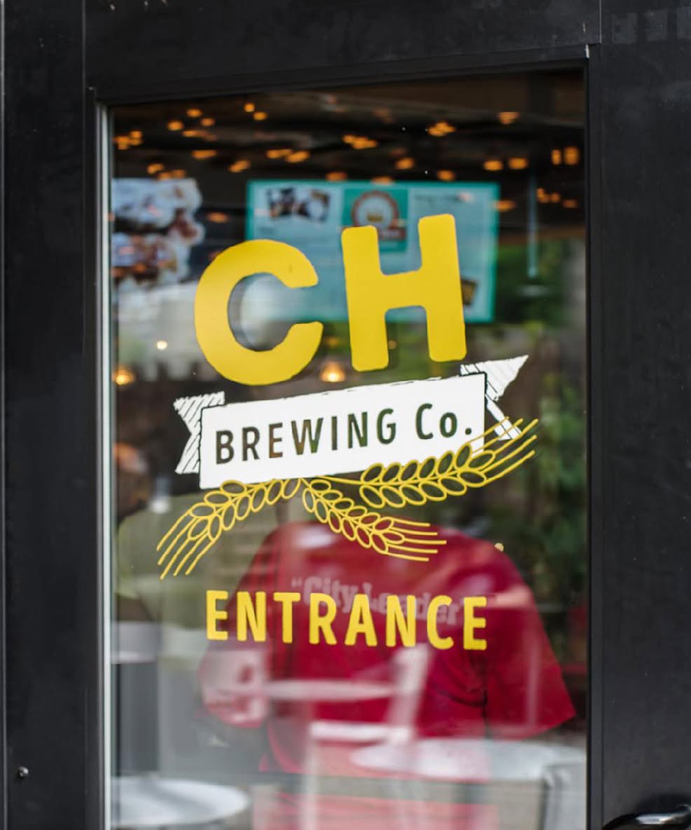 Glass door with "CH Brewing Co. Entrance" in bold yellow letters and a wheat illustration, reflecting a cozy, inviting interior with soft lighting.