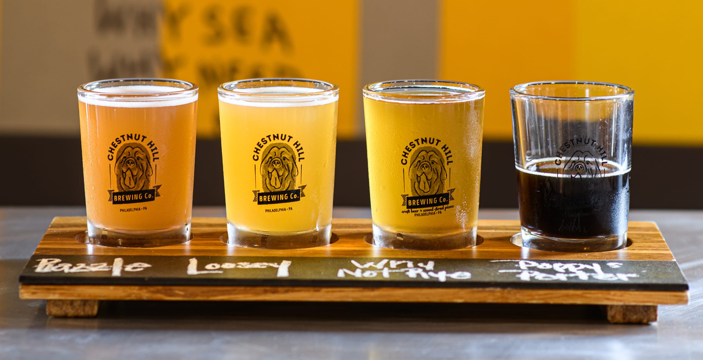 Four beer glasses of varying colors sit on a wooden flight board, labeled "Razzle," "Loosey," "Why Not Rye," and "Poppy Porter" from Chestnut Hill Brewing Co.