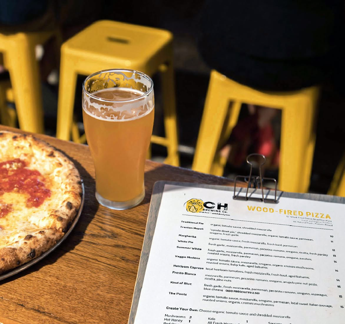 A wooden table with a menu labeled "Wood-Fired Pizza," a glass of pale beer, and a partially visible pizza. Yellow stools are in the background, creating a casual dining atmosphere.