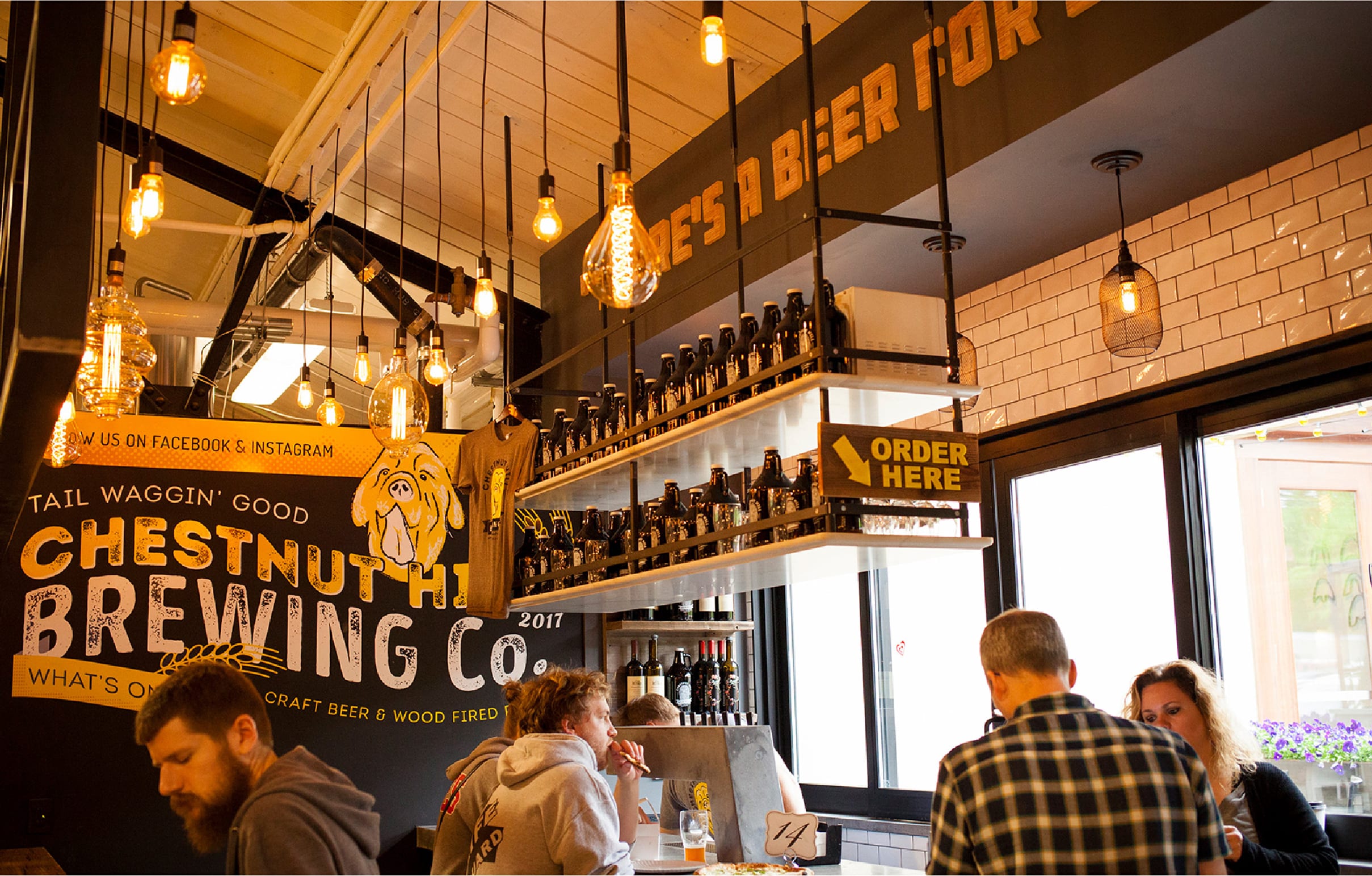A cozy brewery interior with warm lighting, patrons at a counter, and rows of beer bottles. A sign reads "Chestnut Hill Brewing Co." and "Order Here."