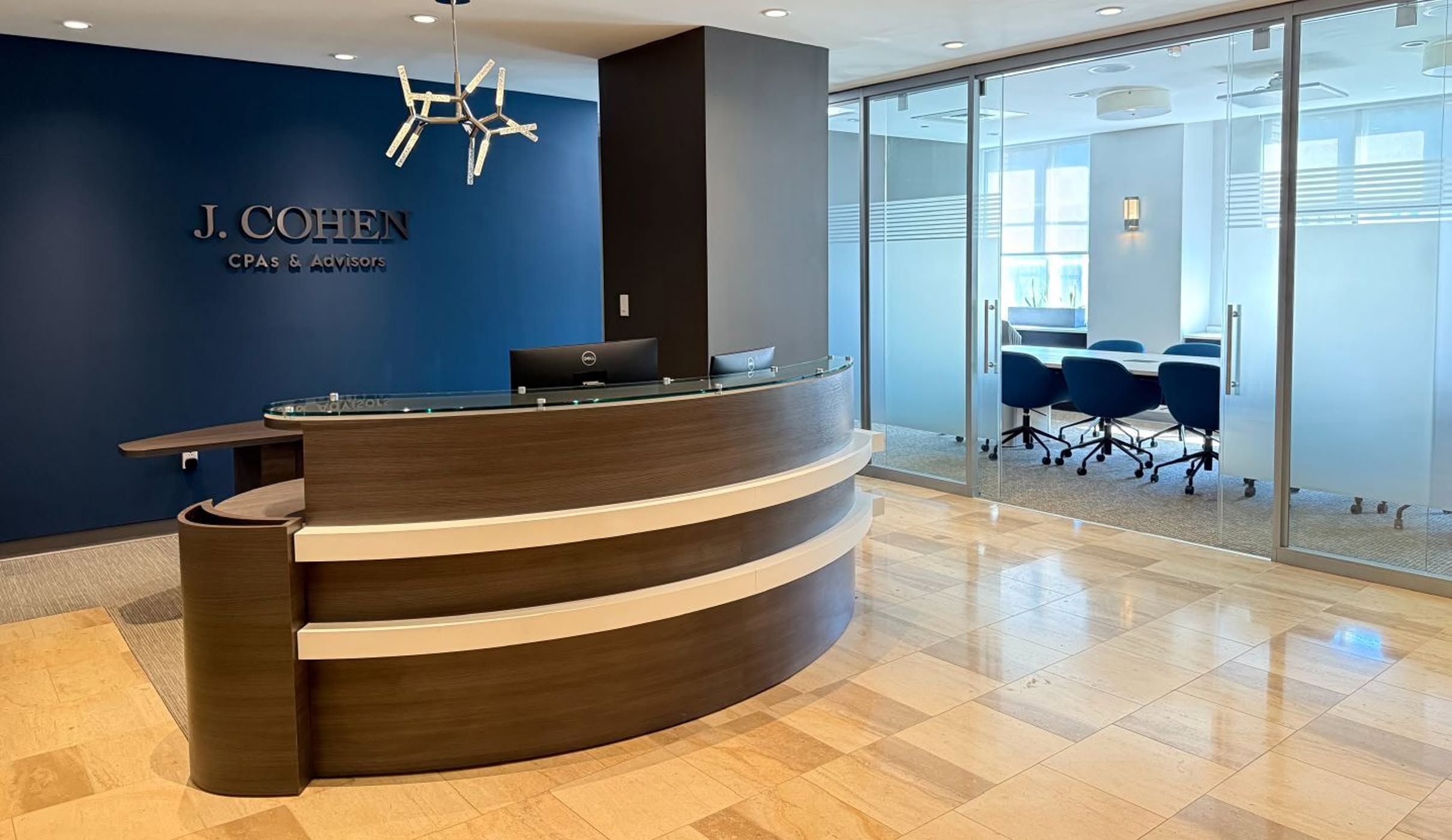 JCohen-Office Modern office reception with a sleek curved desk, dark blue accent wall, and stylish light fixture. The wall has the "J. Cohen CPAs & Advisors" logo. Glass doors reveal a conference room with blue chairs.