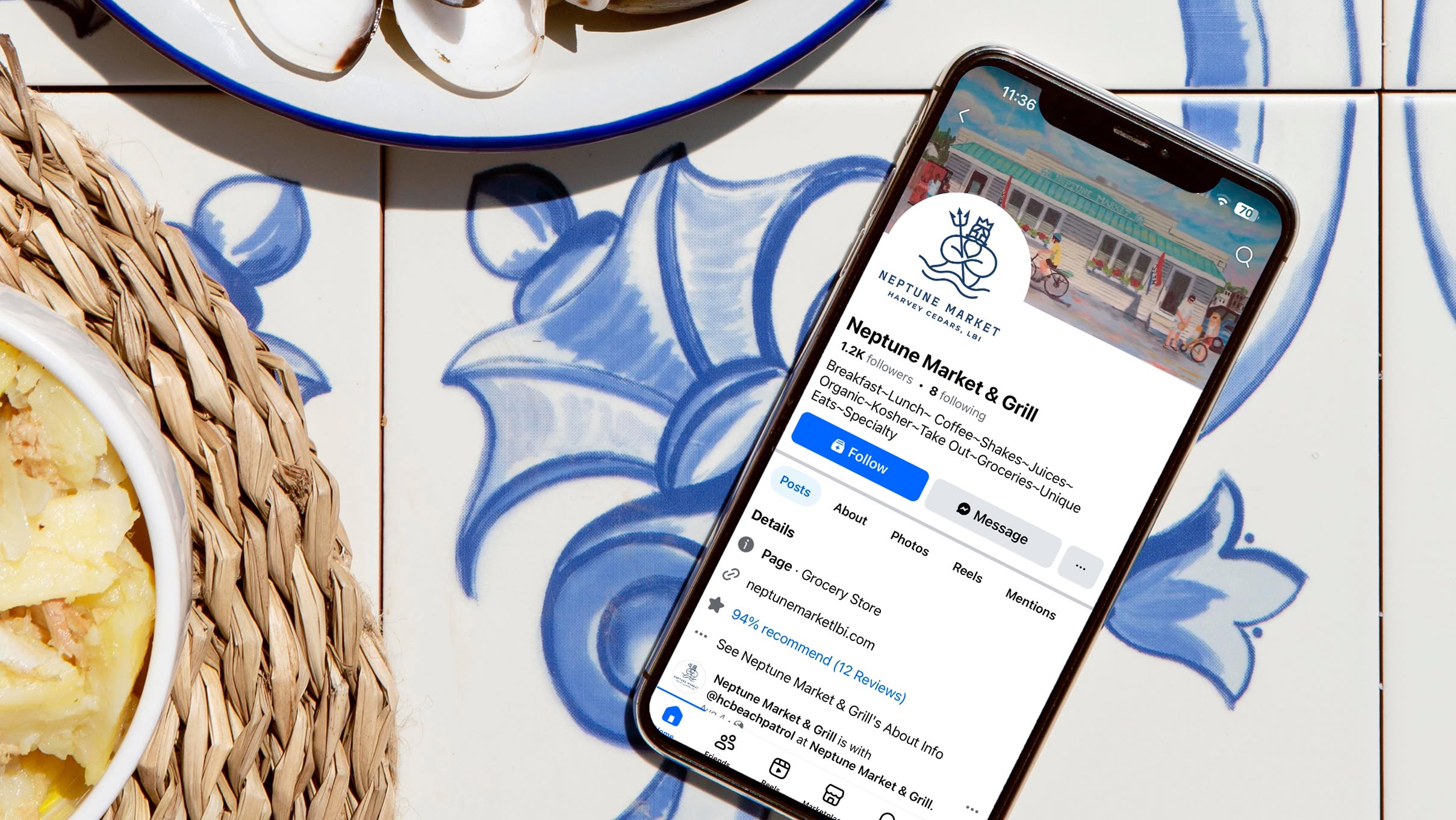 NeptuneMarket-Mobile Smartphone displaying Neptune Market & Grill's profile on social media, placed on a blue floral tiled table. Nearby, dishes with food evoke a casual dining ambiance.