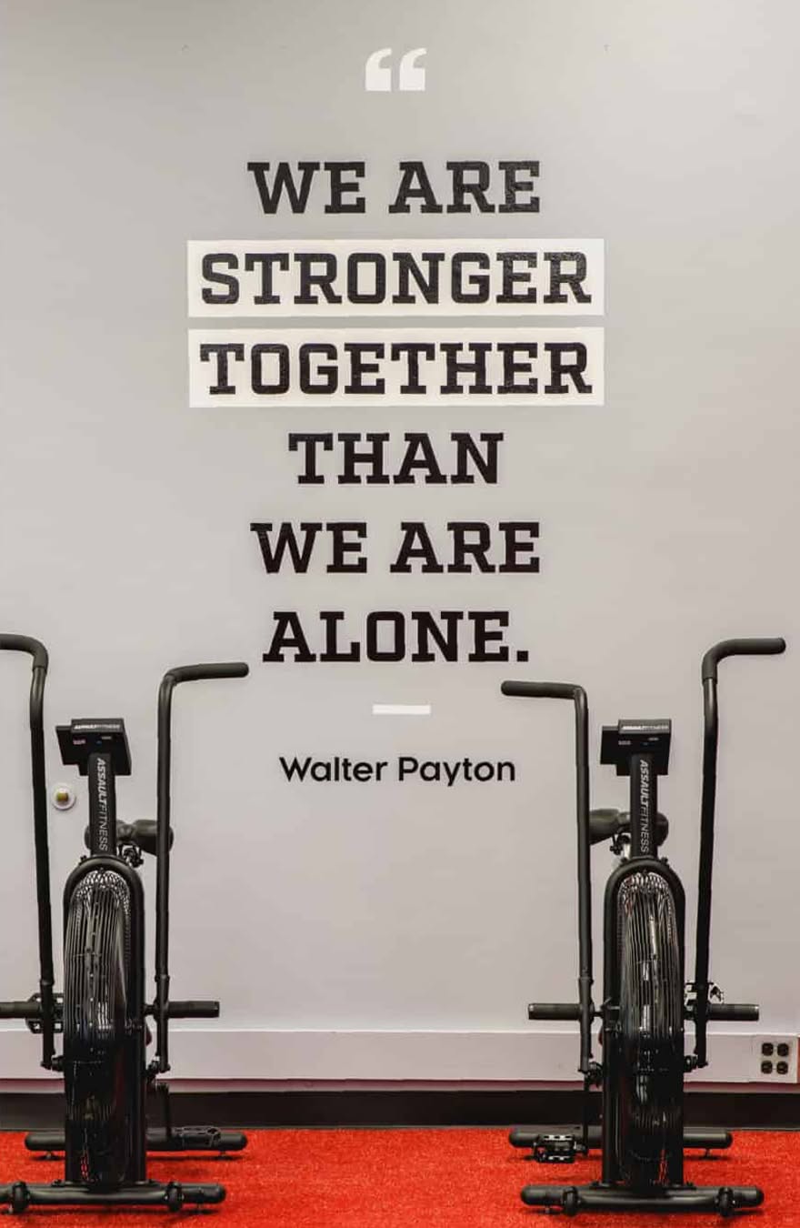 Gym room with red carpet and two exercise bikes in front of a wall quote: "We are stronger together than we are alone." - Walter Payton. Motivational tone.