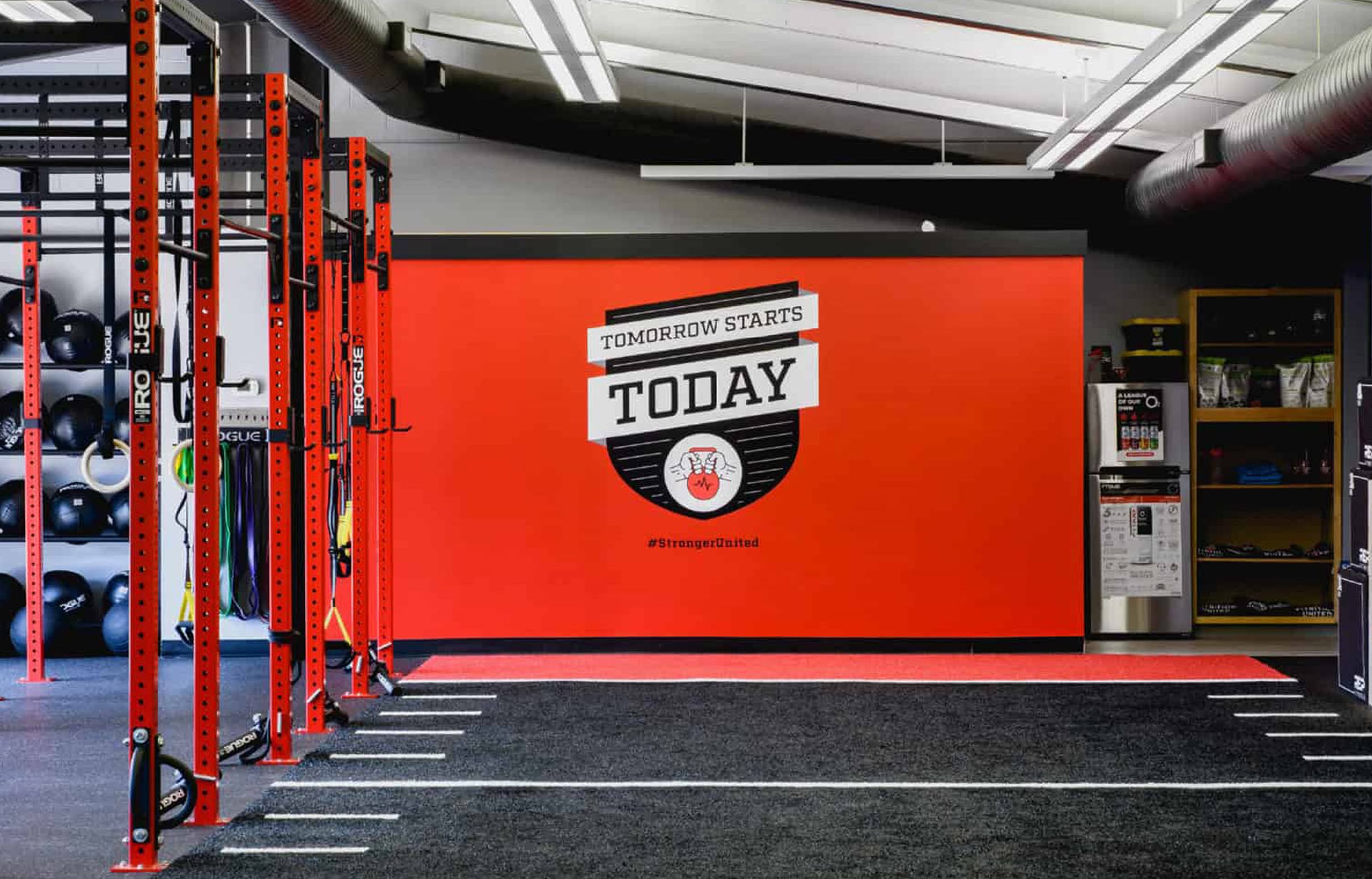A modern gym interior with vibrant red walls displays the motivational text "Tomorrow Starts Today." Red exercise racks line the left, and equipment is neatly organized.