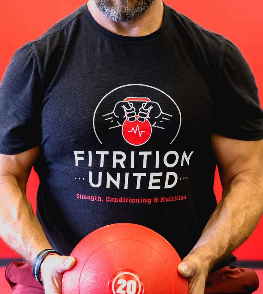 Man in a black "Fitrition United" t-shirt holds a red medicine ball. The shirt features a logo with hands gripping a kettlebell.