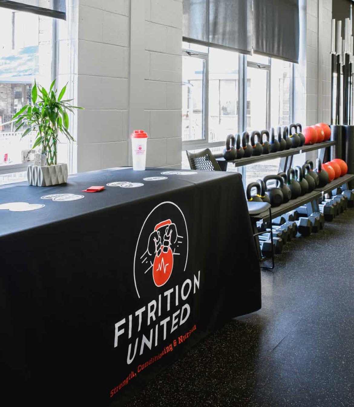 Bright gym room with kettlebells on racks, a table with a "Fitrition United" banner, a plant, coasters, and a shaker bottle, creating a motivational vibe.