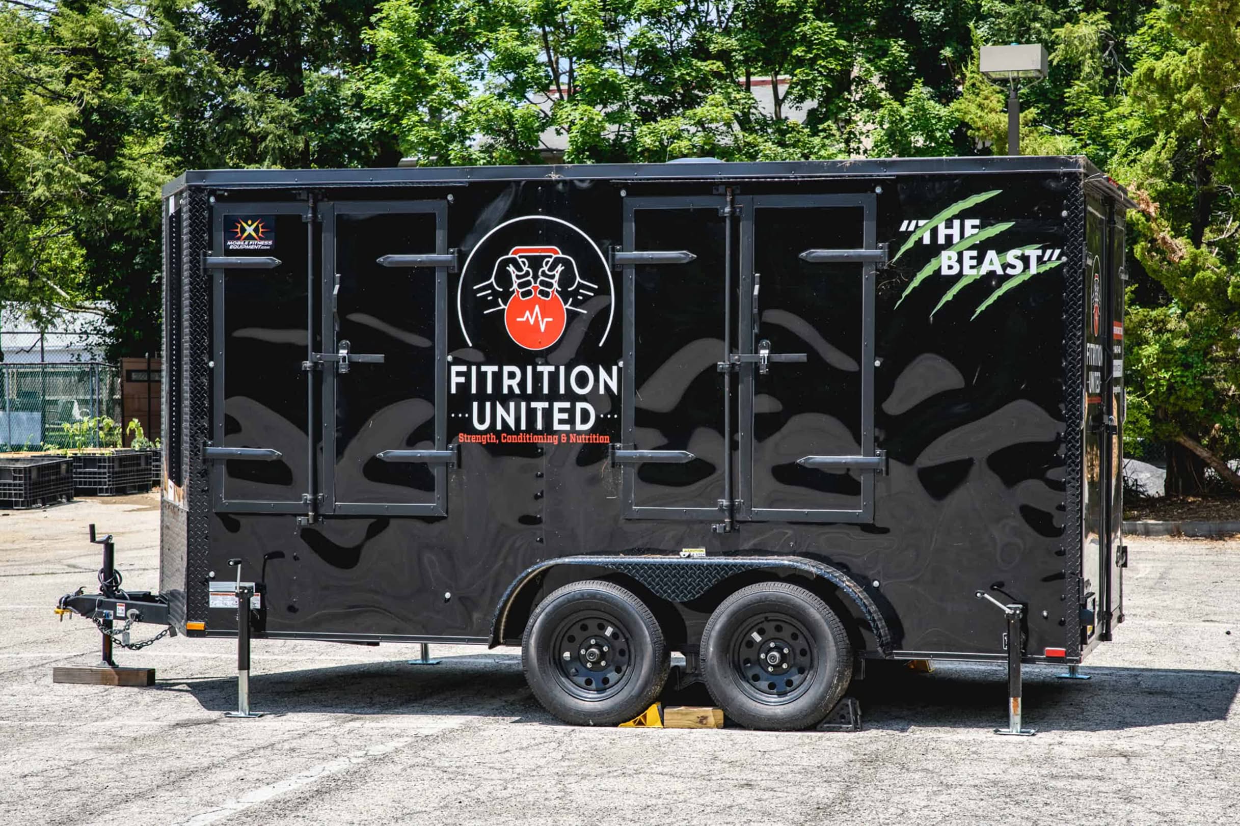 Black trailer labeled "Fitrition United" with a red emblem, parked outdoors. "The Beast" is written in green with claw marks. Trees in background.