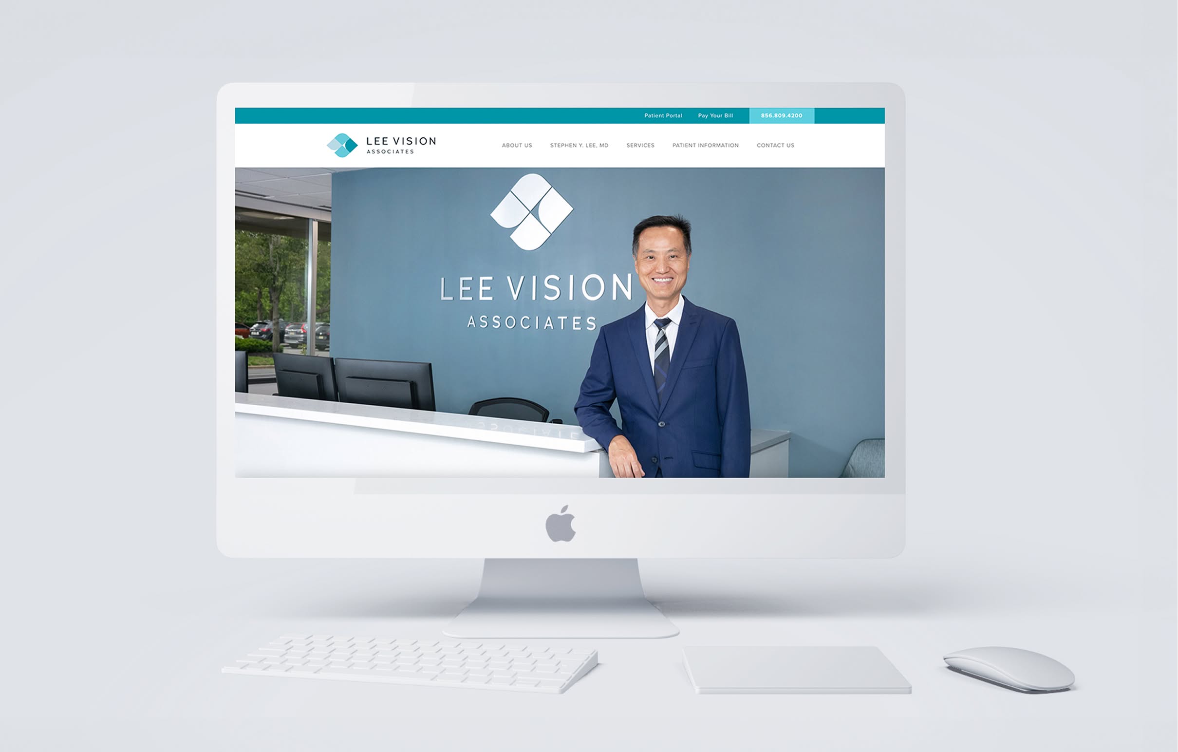 LeeVision-Desktop Smiling man in a suit stands by a reception desk inside a vision care office with "Lee Vision Associates" logo on the wall, viewed on an iMac screen.