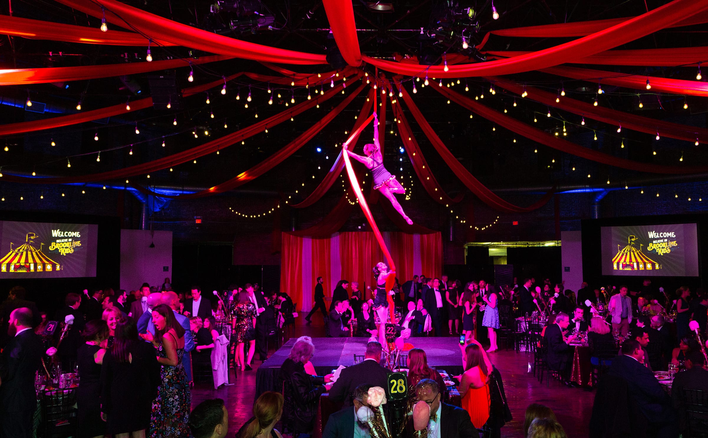 BIBK-Event Aerial performer on red silks above a lively gala, with vibrant string lights and draped fabric overhead. Guests in formal attire socialize below.
