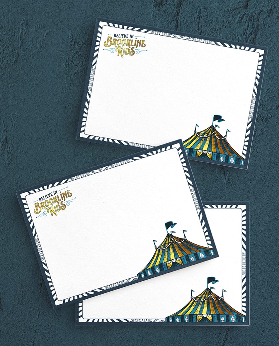 BIBK-Notecards Three white notecards with bold navy borders overlap on a textured blue surface. At the top of the notecards reads "Believe in Brookline Kids" in blue and gold, with a colorful circus tent illustration at the bottom.