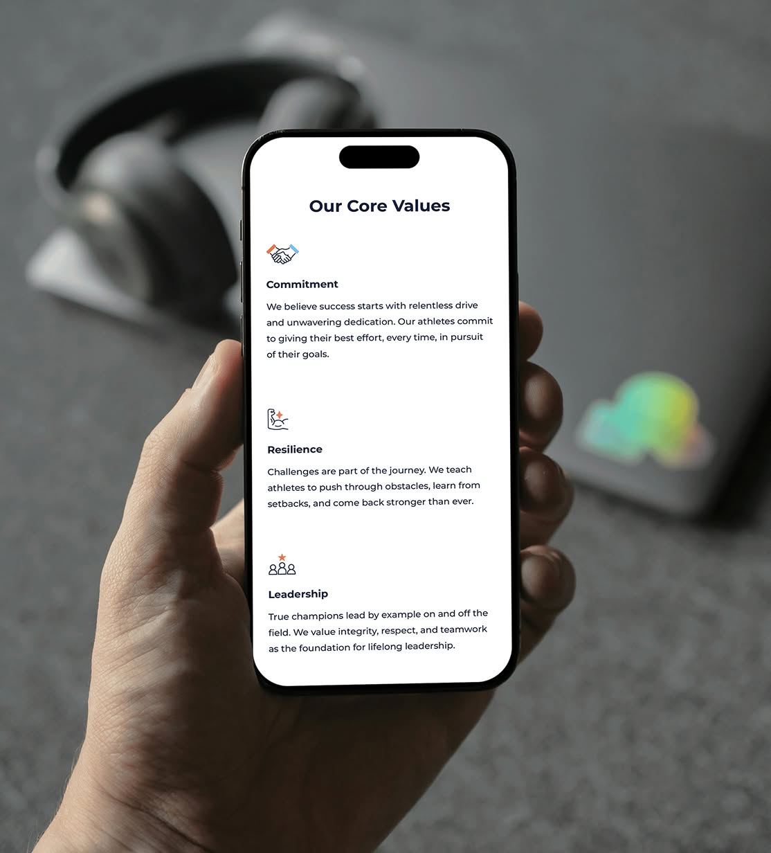 BTS-Phone A hand holds a smartphone displaying a section from the Beyond The Season website. The title "Our Core Values" appears at the top with sections labeled "Commitment," "Resilience," and "Leadership" with accompanying icons. Blurred headphones in the background.