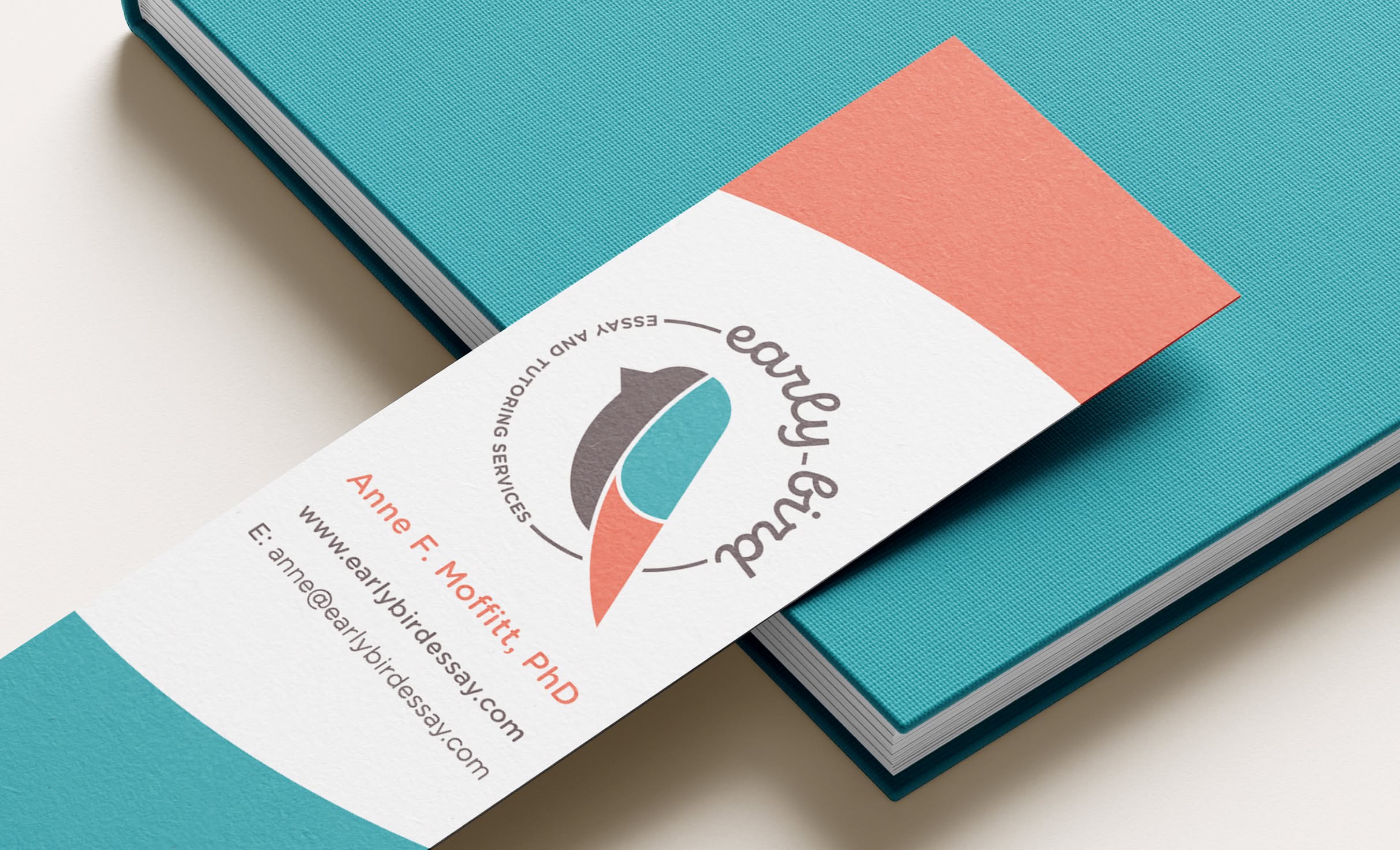 EarlyBird-Bookmark A bookmark rests on top of a teal notebook. The bookmark features color of red and blue blocks at the top and bottom, the Early Bird Tutoring logo in the center, and contact details. The logo includes a stylized bird in brown, blue, and red. In a circle around the bird text reads "early bird" in cursive, with "Essay and Tutoring Services."