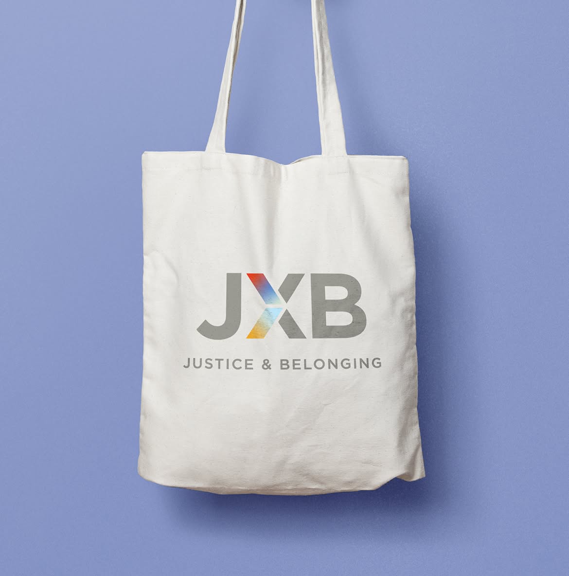 JXB-Bag A white tote bag against a light blue background displays the JXB logo. The logo has the letters "JXB" with a colorful "X" in red, blue, and yellow hues. Below, the text reads "Justice & Belonging."