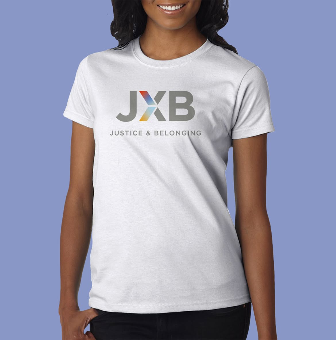 JXB-Shirt A woman wearing a white T-shirt with the JXB logo. The logo has the letters "JXB" with a colorful "X" in red, blue, and yellow hues. Below, the text reads "Justice & Belonging."