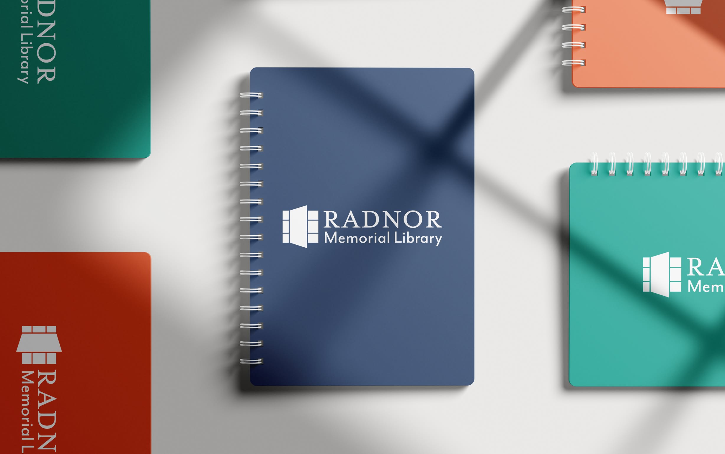 RML-Notebooks A spiral-bound notebook with the Radnor Memorial Library logo on a blue cover is centered. Orange and green notebooks surround it on a light background.